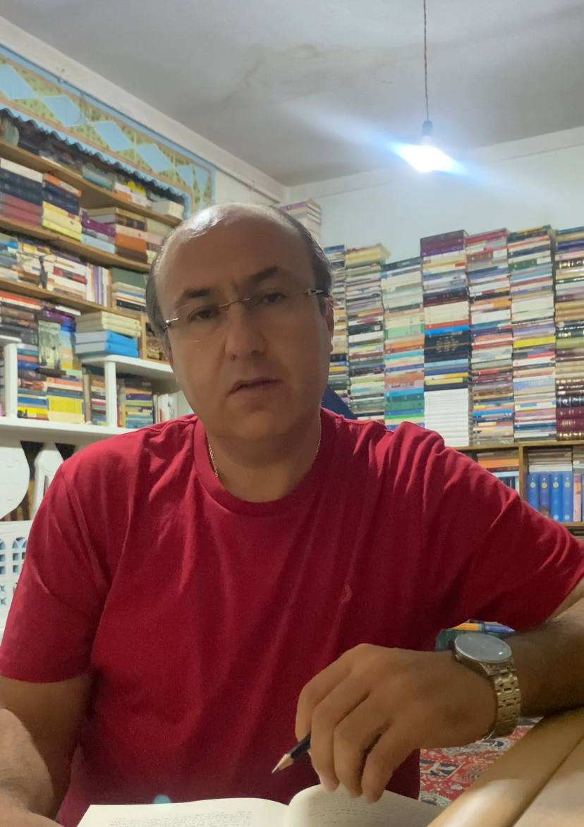 Image of Iranian poet Alireza Nouri, middle aged man, bald and wearing glasses in a red t-shirt looking studious in a room full of books.