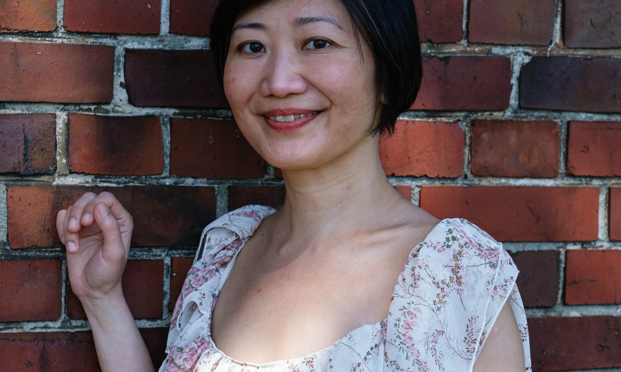 Translator Christina Ng smiles in a white floral dress, standing with one hand raised against a red brick wall.