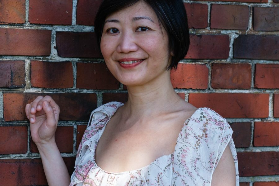 Translator Christina Ng smiles in a white floral dress, standing with one hand raised against a red brick wall.